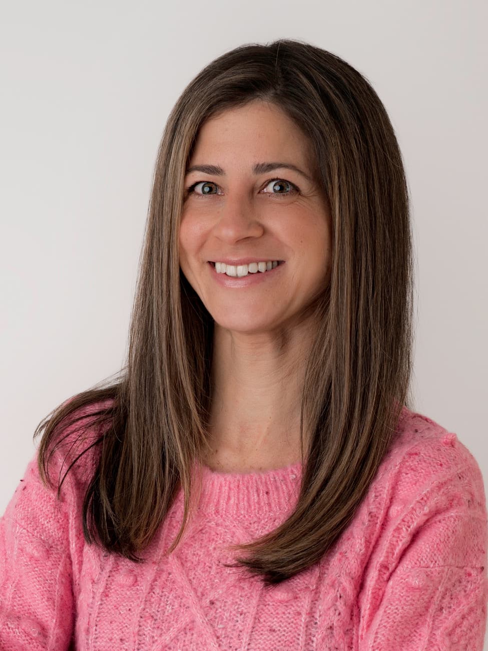 Smiling woman with long brown hair wearing a pink textured sweater on a white background.